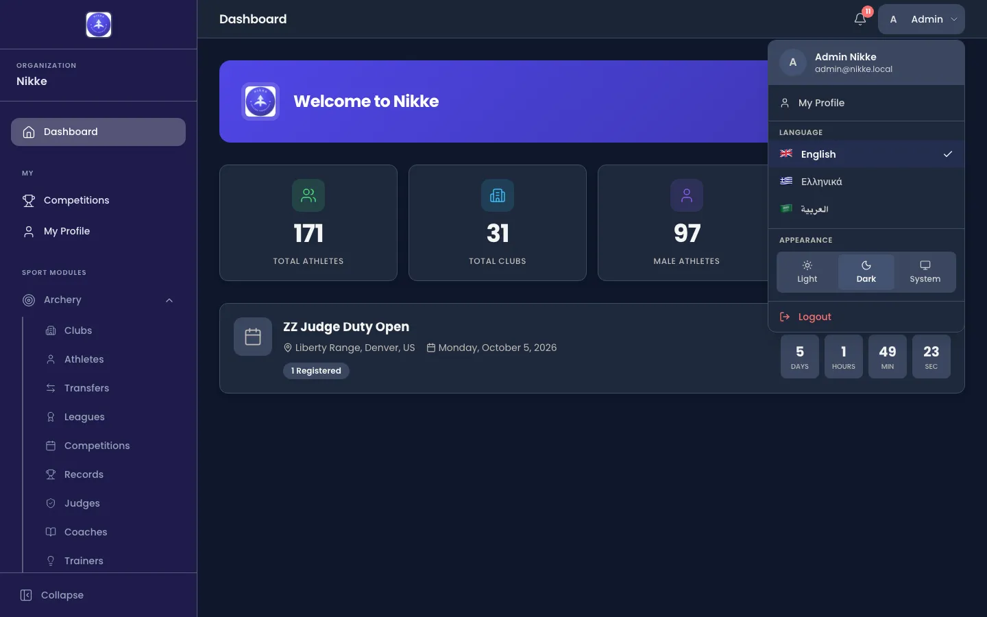 The Velos account menu open over the dashboard, showing the signed-in person's full name with their email address beneath it, a My Profile item, a Language section listing English, Greek and Arabic, an Appearance section with Light, Dark and System buttons, and a Logout item.
