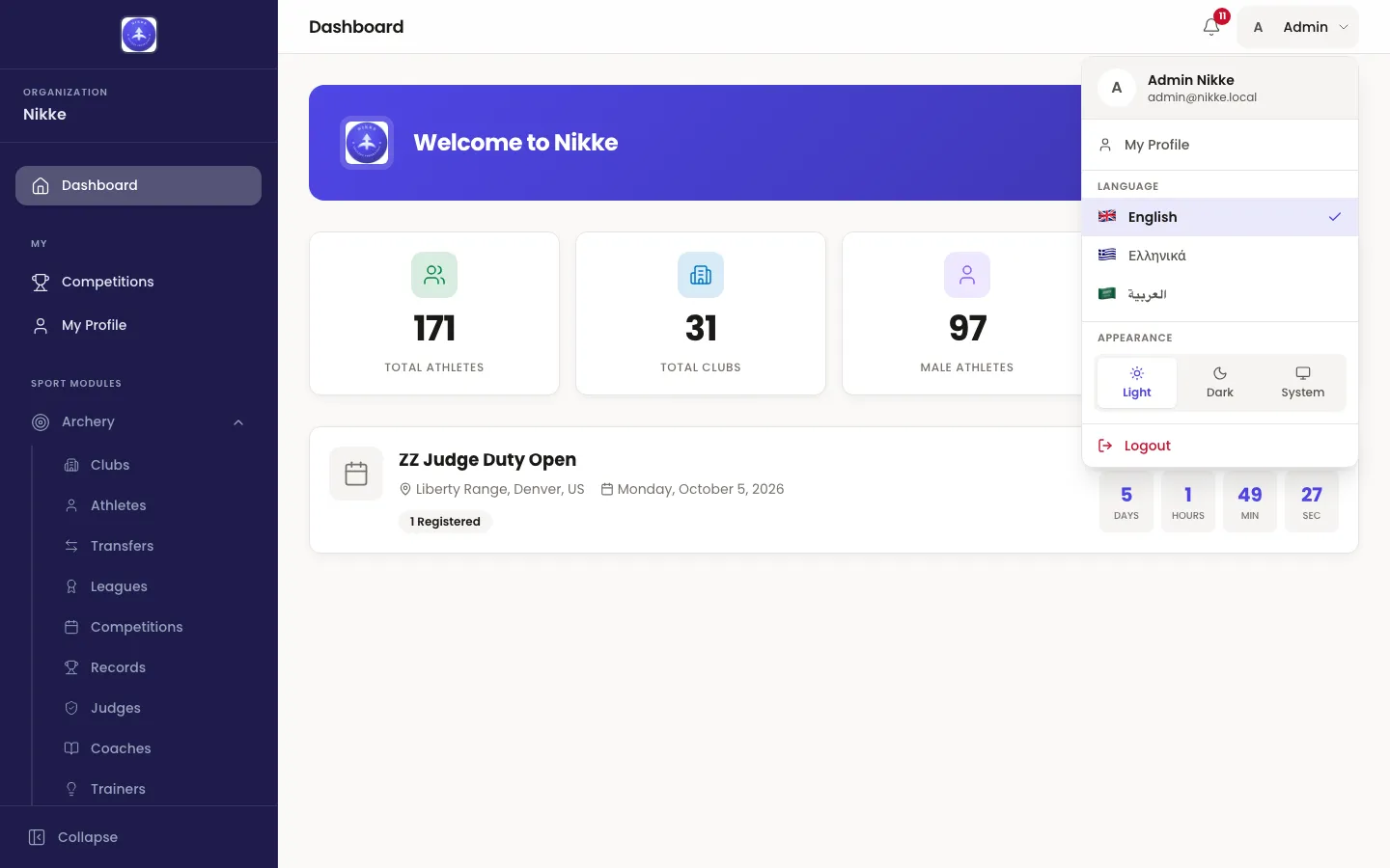 The Velos account menu open over the dashboard, showing the signed-in person's full name with their email address beneath it, a My Profile item, a Language section listing English, Greek and Arabic, an Appearance section with Light, Dark and System buttons, and a Logout item.